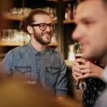 Handsome smiling hipster man sitting at pub near bar counter, holding pint of beer and listening interesting stories. Nightlife.