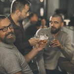Homme joyeux et barbu tenant un verre de whisky en cristal, regardant la caméra et souriant. Clients du bar assis au comptoir derrière, communiquant et buvant de l'alcool d'élite.